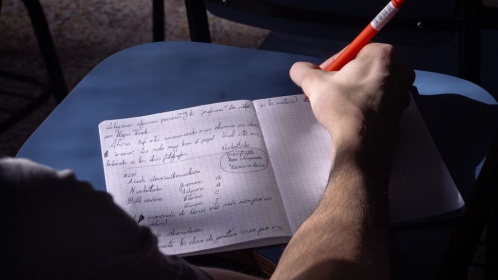 Mano escribiendo con lápiz sobre cuaderno y libros de fondo